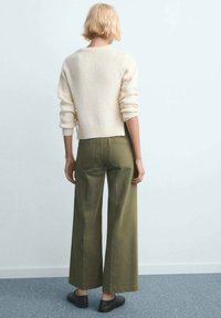 Maglione lavorato a maglia color crema, pantaloni a gamba larga oliva, scarpe nere. Il maglione ha polsini e orlo a coste; i pantaloni presentano tasche posteriori e vestibilità ampia.