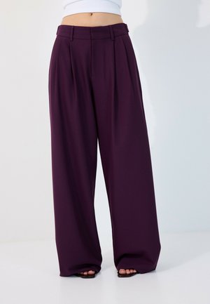 Pantalon large taille haute violet foncé porté avec des chaussures noires à bouts ouverts et un top court blanc sur un fond uni.