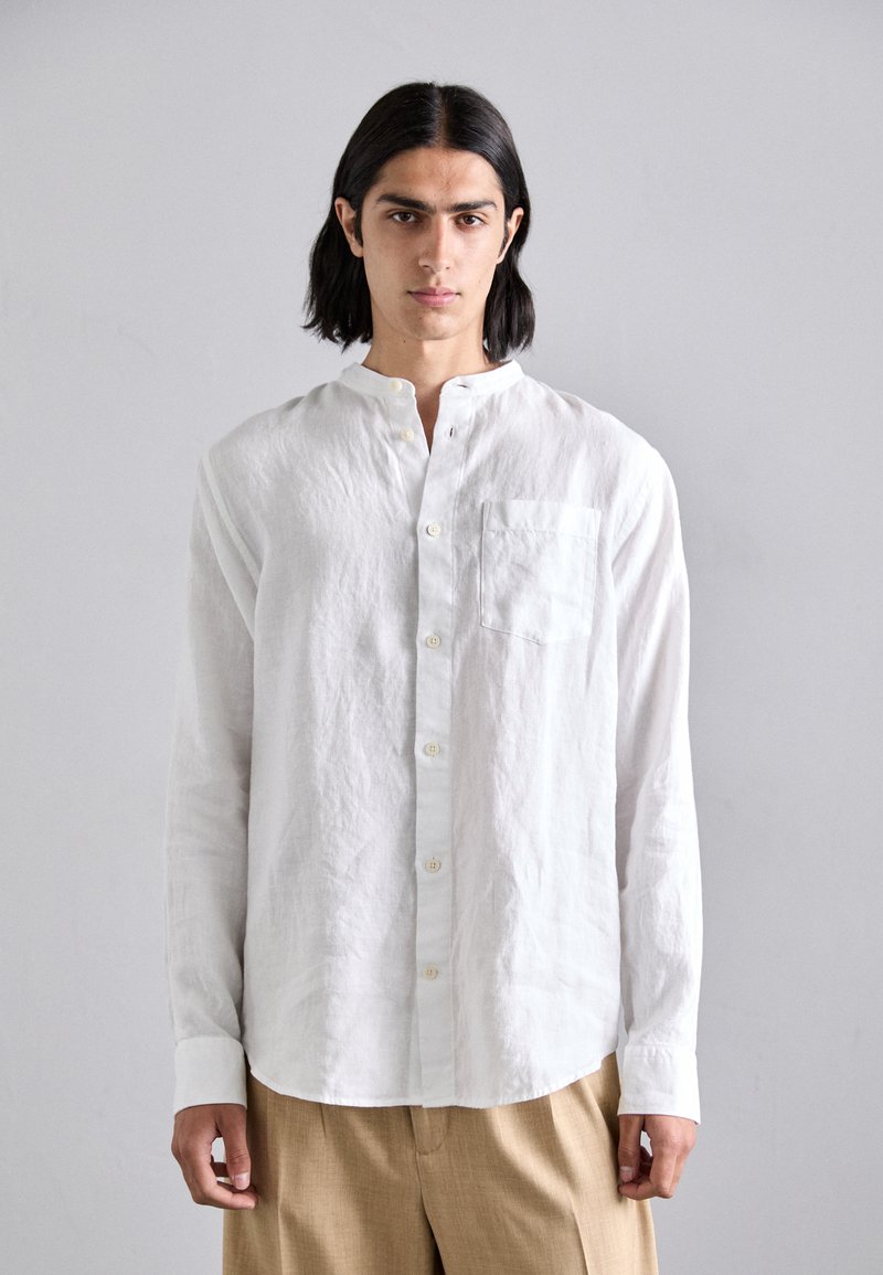 Camicia bianca in lino a maniche lunghe con colletto alla cinese, una sottile texture e taschino frontale. Presenta una chiusura a pulsante e un taglio rilassato.