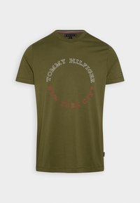 Camiseta de algodón verde oliva con un logo redondo que presenta "TOMMY HILFIGER" y "NEW YORK CITY" en texto blanco y rojo en contraste. Mangas cortas.