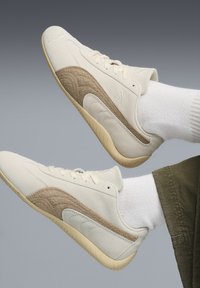 Białe sneakersy z beżowymi akcentami, zamszowymi i teksturowanymi panelami, płaską gumową podeszwą oraz minimalistycznym wzornictwem. Zawierają branding po boku.