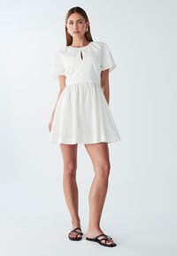 Biele krátke šaty s nafúknutými rukávmi, okrúhlym výstrihom a detaily v tvare kľúčovej dierky. A-liniová silueta, ľahký materiál, doplnené čiernymi sandálmi.