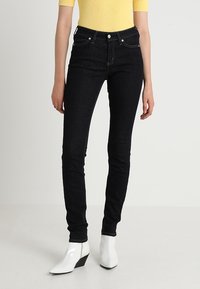 Jeans de mezclilla negros de tiro alto con un ajuste ajustado, combinados con una blusa amarilla de canalé y botas blancas de tacón angulado, exhibidos sobre un fondo neutro.