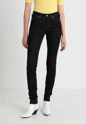 Jeans de mezclilla negros de tiro alto con un ajuste ajustado, combinados con una blusa amarilla de canalé y botas blancas de tacón angulado, exhibidos sobre un fondo neutro.
