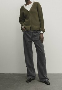 Persona che indossa un cardigan a coste verde oliva sopra una camicia bianca, abbinato a jeans scuri grigi larghi e scarpe nere, in piedi all'interno.