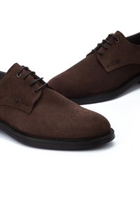 Zapatos de vestir de gamuza marrón con una textura suave, presentan un diseño de punta afilada, cordones negros y un sutil detalle de marca en el costado.