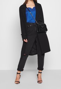 Musta pitkä takki, sininen satiininen camisole pitsihelman kanssa, mustat korkeavyötäröiset farkut kulutuksella, pieni musta käsilaukku ja remmisandaalit.