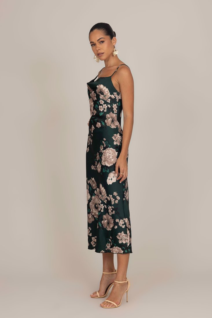 Robe en satin vert à fleurs avec un corsage ajusté, des fines bretelles et une longueur mi-mollet. Présente de grandes fleurs roses et blanches, portée avec des talons nude.