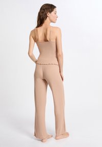 Donna in piedi, scalza, indossa un top beige a coste senza maniche e pantaloni larghi coordinati, girata di spalle su uno sfondo bianco uniforme.
