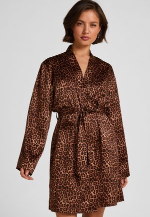 Frau trägt ein braun-schwarz gemustertes Satin-Wickelkleid mit Leopardenmotiv, langen Ärmeln und einem gebundenen Taillengürtel, vor einem schlichten grauen Hintergrund.