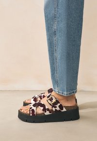 Platvormsandaalid, millel on pruunide ja valgete lehmakujutistega mustrid, pandlaga ning risti rihmadega, kantud siniste teksapükstega.