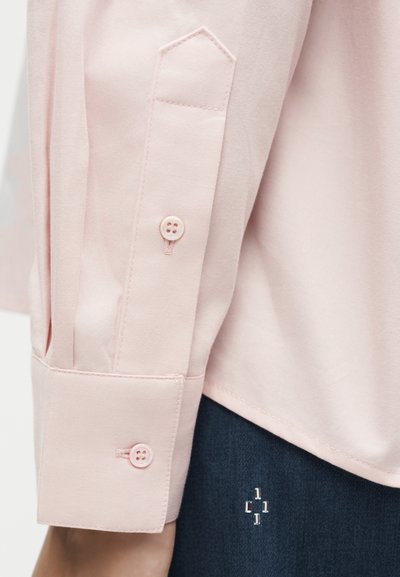 Κοντινή λήψη μανικετιού ροζ πουκαμίσου με δύο κουμπιά, φορεμένο πάνω από σκούρο μπλε παντελόνι με μικρές μεταλλικές διακοσμήσεις.