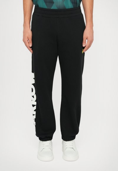 Schwarze Jogginghosen mit elastischem Bund, tapered Cuffs, großen weißen Buchstaben an der Seite und einem kleinen grünen Logodetail.