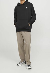 Felpa nera con cappuccio, tasca frontale e un piccolo logo bianco sul petto, abbinata a pantaloni beige chiaro e scarpe da ginnastica con motivo.