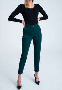 Zielone, dopasowane spodnie o slim fit, z przednimi kieszeniami, kontrastowymi szwami i pojedynczym zapięciem na guzik, połączone z czarną bluzką z długim rękawem.