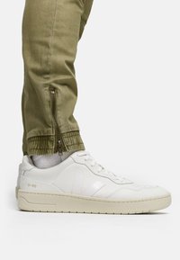 Białe skórzane sneakersy o gładkim wykończeniu, z teksturowaną podeszwą i subtelnym detalem logo. Połączone z oliwkowymi zwężanymi joggerami z zamkami.