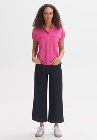 Camicia button-up rosa a maniche corte con due tasche sul petto, abbinata a jeans a gamba larga scuri e sneakers bianche. Tessuto leggero e morbido.