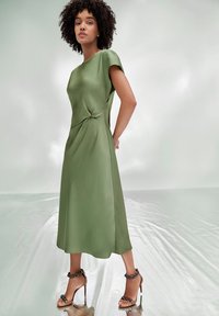 Robe en satin vert à manches courtes, avec un détail noué à la taille et une silhouette évasée fluide, accompagnée de talons décorés à lanières.