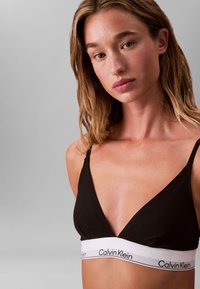 Young woman with light brown hair wears a black Calvin Klein bralette with white elastic band against a plain gray background.