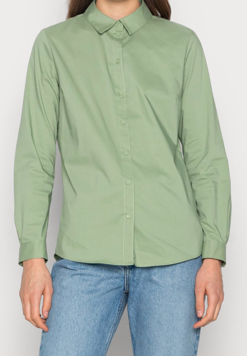 Ljusgrön långärmad skjorta med krage och knäppning, tillverkad av ett mjukt material. Kombinerad med ljusblå denimjeans.