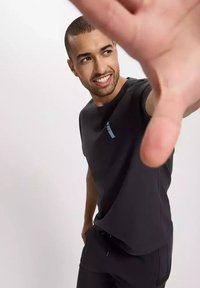 Czarna bawełniana koszulka z okrągłym dekoltem, z niebieskim logo na lewej piersi. Model pozuje z ręką wyciągniętą przed siebie.