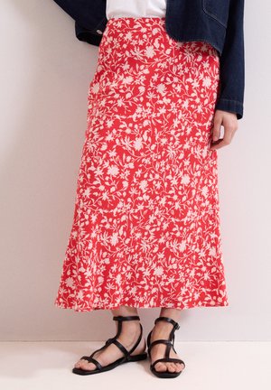 Persoon draagt een rode midi rok met bloemenprint, zwarte platte sandalen met bandjes, een wit topje en een donkerblauwe spijkerjas, staand tegen een lichte muur.