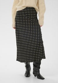 Jupe midi noire à pois crème, en tissu lisse, avec une légère évasure et une fente latérale. Associée à des bottes noires montant jusqu'aux genoux.