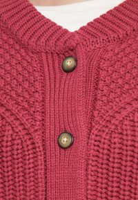 Κοντινή λήψη ενός κόκκινου πλεκτού ζακέτας με υφές και δύο καφέ κουμπιά πάνω από ένα λευκό πουκάμισο.