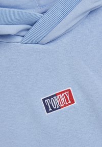 Světle modrá mikina s kapucí z měkké látky, která má vyšívané logo v červené a námořnické modré barvě. Žebrované manžety a detail kolem krku zlepšují design.