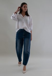 Vrouw die haar aanpast, gekleed in een witte blouse, blauwe jeans en beige hakken, tegen een effen grijze achtergrond.