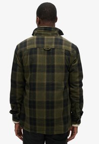 Camisa de franela a cuadros verdes y negros con un cuello alto, puños con botones y un detalle trasero para colgar. Textura suave y corte relajado.