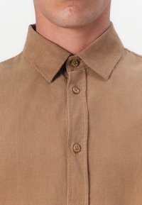 Chemise en velours côtelé beige avec un col pointu, présentant des fermetures à boutons, un tissu texturé et un motif côtelé sur le devant.