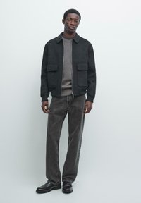 Giacca grigia simile a lana con colletto e due tasche frontali, indossata sopra un maglione grigio e jeans grigio scuro, completata da scarpe nere.