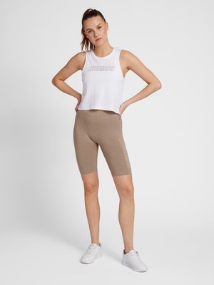 Kobieta ubrana w biały bezrękawnik z napisem „HUMMEL”, beżowe krótkie spodenki rowerowe z napisem z boku i białe trampki, stojąca z rękami na biodrach.