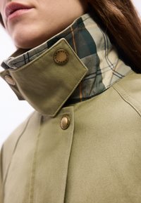 Κοντινό πλάνο ενός ατόμου που φοράει μπεζ μπουφάν με ανασηκωμένο γιακά, αποκαλύπτοντας πράσινη και μπεζ καρό φόδρα και χάλκινους κουμπωτούς κουμπίες.