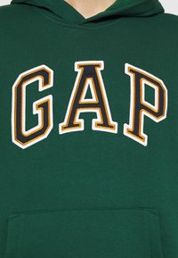 Felpa verde con tasca a marsupio, caratterizzata dal logo ricamato "GAP" in bianco e arancione. Tessuto morbido, finitura testurizzata, vestibilità rilassata.