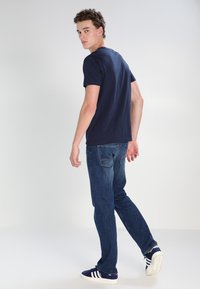 Navy blue cotton t-shirt paired with dark blue jeans. Model facing sideways, wearing white and navy sneakers with three stripes. Simple design.