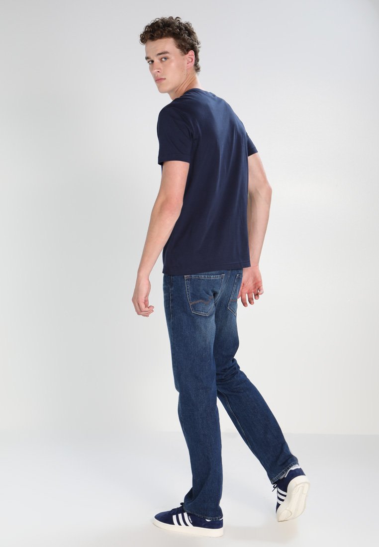 Navyblaues Baumwoll-T-Shirt kombiniert mit dunkelblauen Jeans. Das Modell steht seitlich, trägt weiße und navyblaue Turnschuhe mit drei Streifen. Einfaches Design.