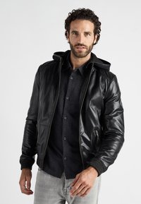 Schwarze Lederjacke mit Kapuze, Reißverschlüssen und gerippten Strickbündchen, getragen über einem dunklen Hemd mit Knöpfen, kombiniert mit hellgrauen Jeans.