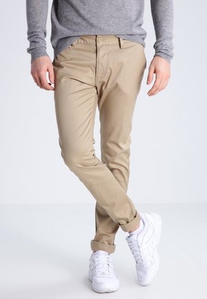 Man draagt beige opgerolde broek, witte sneakers en een grijze trui met lange mouwen, staat met gekruiste benen.