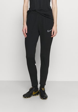 Personne portant un pantalon de survêtement Nike noir avec un logo blanc sur la cuisse et des chaussures de football noires avec un swoosh doré, debout devant un fond uni.