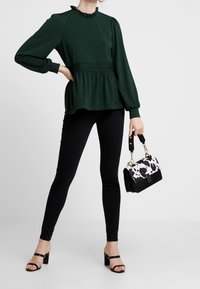 Tmavě zelená peplum blůza s volánovým límcem a krajkovými detaily, kombinovaná s černými džíny a kabelkou s motivem krávy. Černé sandály na vysokém podpatku.