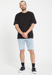 Sort bomulds T-shirt med lille rødt logo, parret med lyseblå denimshorts. Båret med sorte sneakers og sokker, stående i en neutral pose.