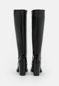 Bottes noires en cuir hautes jusqu'au genou avec une texture lisse, un talon épais et haut, et une fermeture éclair à l'arrière, avec un accent en boucle au niveau du talon.