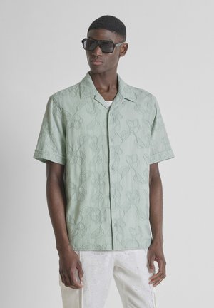 Mann mit übergroßer schwarzer Sonnenbrille, hellgrüner kurzärmeliger Bluse mit Blumenstickerei und weißen Hosen vor schlichtem Hintergrund.