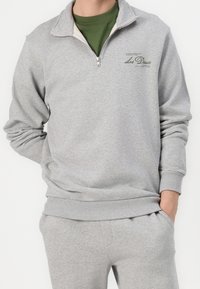 Sweat-shirt gris à fermeture éclair avec un col montant, fabriqué en tissu doux. Présente un petit logo vert sur la poitrine ainsi que des poignets et un ourlet côtelés.