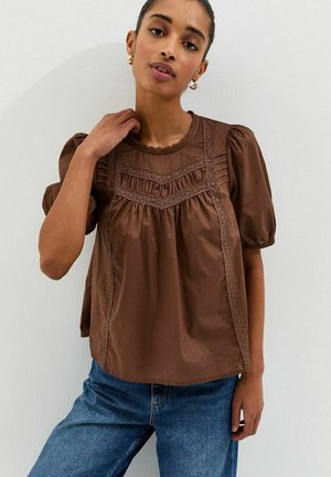 Femme portant un chemisier marron à manches bouffantes avec des détails en dentelle et un jean bleu, posant devant un mur blanc uni.