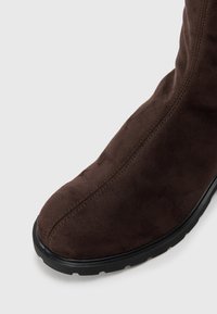 Botte en daim marron foncé avec des coutures visibles et une semelle en caoutchouc noir, présentée en gros plan sous un angle montrant l'embout et le côté supérieur.