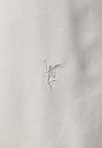 Vaaleanharmaa kangas, jossa on pehmeä ja sileä pinta, ja hienovarainen valkoinen kirjailtu keijukaiskuvio tanssiposeerissa.