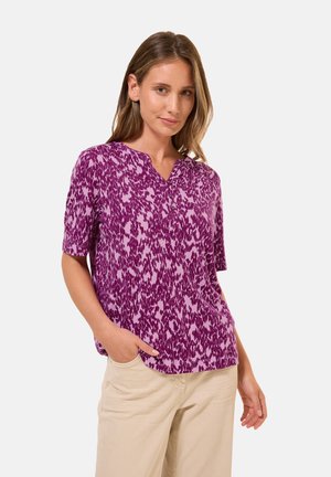 Blouse violette à manches courtes avec motif abstrait, décolleté en V et tissu léger. Offre une coupe décontractée et des poches sur les côtés.
